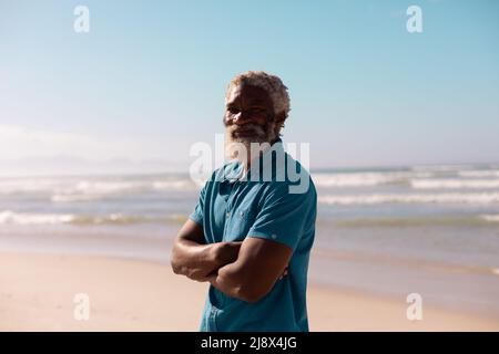 Porträt eines bärtigen afroamerikanischen Senioren mit gekreuzten Armen, der vor dem Meer und dem blauen Himmel steht Stockfoto