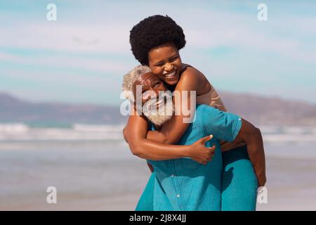 Verspielte bärtige afroamerikanische Senioren huckbacking glückliche reife Frau gegen Meer und Himmel Stockfoto