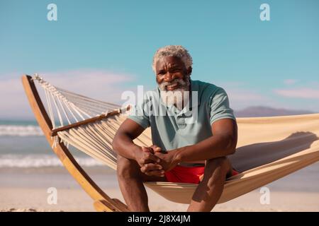 Porträt eines glücklichen afroamerikanischen bärtigen älteren Mannes, der im Sommer in einer Hängematte am blauen Himmel sitzt Stockfoto