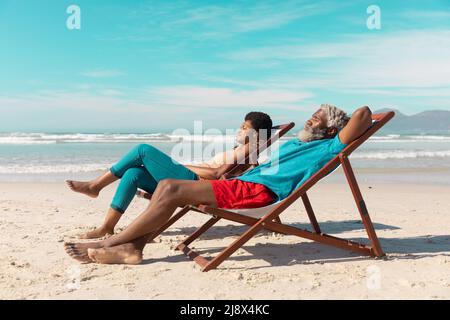 Seitenansicht eines afroamerikanischen Paares, das sich im Sommer auf Liegestühlen vor dem Meer und dem blauen Himmel entspannen kann Stockfoto