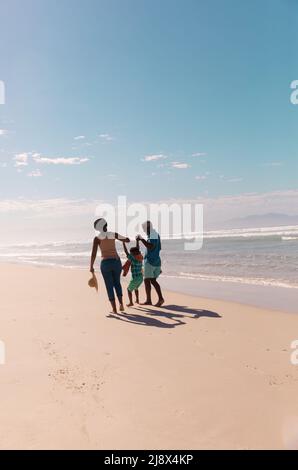 afroamerikanische Großeltern halten die Hände des Enkels und gehen am Sandstrand gegen den blauen Himmel Stockfoto