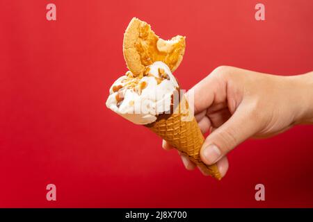 Hand hält einen frischen Eiskegel mit einem Stück Keks mit einem Bissen auf Rot Stockfoto