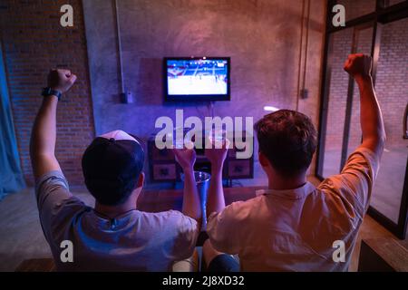Zwei männliche Freunde, die ein Bierglas halten, feiern zu Hause Sport im Fernsehen. Stockfoto