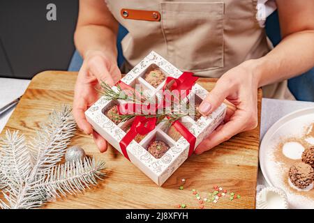 Eine Konditorin hält Schokolade in Schachteln. Hochwertige handgefertigte Schokoladentrüffel. Süßes Geschenk zu Weihnachten oder Neujahr. Nahaufnahme der weiblichen Hände und t Stockfoto