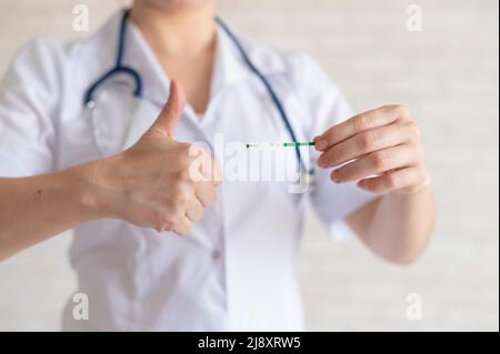 Ein gesichtsloser Arzt in einem medizinischen Mantel und mit einem Stethoskop hält einen positiven Schwangerschaftstest ab. Der Gynäkologe zeigt einen Daumen nach oben. Stockfoto