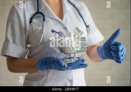 Ein gesichtsloser Arzt mit einem medizinischen Mantel und einem Stethoskop hält einen kleinen Einkaufswagen voller Medikamente. Apothekenkonzept. Der Apotheker zeigt einen Daumen Stockfoto