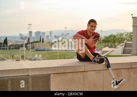 Junger Mann mit einer Behinderung, der ein Mobiltelefon benutzt, während er sich im Freien entspannt. Stockfoto