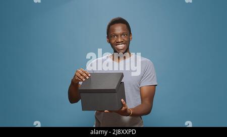 Fröhlicher Mann, der die Geschenkbox öffnet, um den Kauf vor der Kamera zu sehen und den Pappeinkauf auszupacken. Person lächelt und entpackt Lieferung Paket aus E-Commerce-Geschäft. Stockfoto