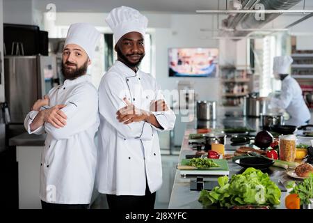 Multiethnische männliche Köche stehen in der professionellen Küche des Restaurants mit gekreuzten Armen, während sie Zutaten für das Gericht zubereiten. Kulinarische Experten tragen Kochhüte, während sie in der Nähe der Kochstation stehen. Stockfoto