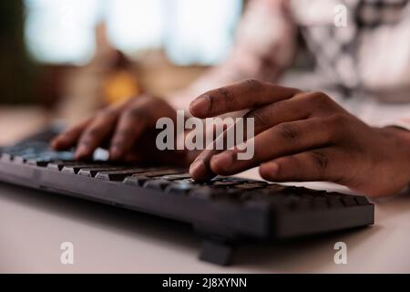 Nahaufnahme des afrikanischen amerikanischen männlichen Content Creator, der Post für soziale Medien auf der Computertastatur im Wohnzimmer schreibt. Selektiver Fokus auf Mann Hände arbeiten Remote Typing Blog-Artikel. Stockfoto