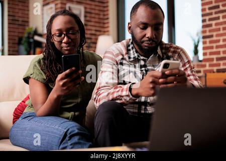 Freund ignoriert verärgert Freundin nach Streit verbringen Zeit auf Smartphones sitzen auf der Couch. afroamerikanisches Paar, das zusammen lebt, hat Probleme, nicht miteinander im Wohnzimmer zu sprechen. Stockfoto