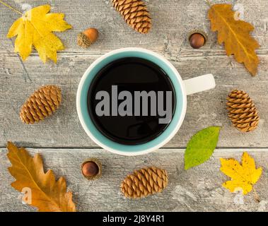 Draufsicht auf einen Becher mit schwarzem Kaffee, Herbstblättern, Kiefernholzkännern und Eicheln auf verwittertem Holzpaneelhintergrund. Stockfoto