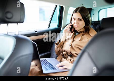 Geschäftsfrau mit Laptop empfangen von Anrufen auf dem Rücksitz eines Autos. Stockfoto