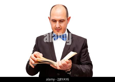 Ein kahlköpfiger, erwachsener Mann aus dem Kaukasus, der in einem schwarzen Anzug, einem weißen Hemd und einer blauen Fliege gekleidet ist, liest ein Buch. Der Hintergrund ist weiß. Stockfoto