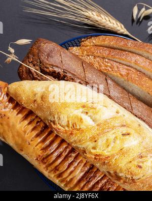 Verschiedene Arten von Naturbroten. Frische Brotscheiben im blauen Korb mit Roggen- und Weizenohren auf schwarzem Hintergrund. Knusprige französische Baguettes. Stockfoto