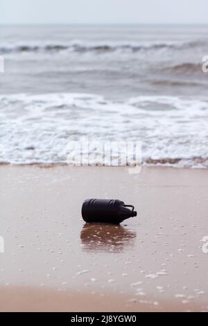 Eine leere schwarze Plastikflasche am Strand, die von der Meeresströmung in den Sand gebracht wird. Konzept der Wasser- und Meeresverschmutzung. Vertikale Zusammensetzung Stockfoto