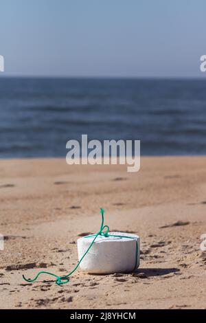 Ein geformtes Stück weißem Styropor mit einer grünen Schnur um ihn herum, das auf dem Sand in der Nähe des Ozeans am Strand steht. Konzept der Meeresverschmutzung Stockfoto