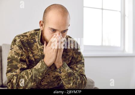 Deprimierter junger Soldat, der an PTSD leidet, sitzt auf dem Sofa und denkt an Krieg Stockfoto