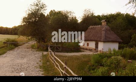 Kleines niedliches Haus, umgeben von einem einfachen Holzzaun in ländlicher Umgebung. Zwei kleine Fenster im Haus. Das Dach ist mit Strohdach versehen. Dichter Wald in der Nähe des Hauses. Stockfoto