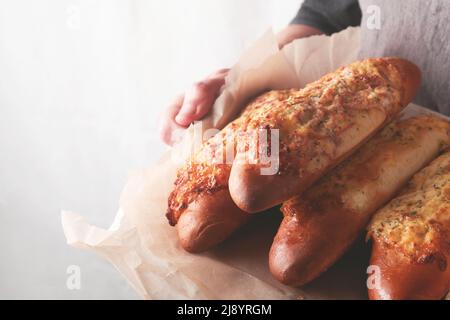 Hände eines Mannes, der ein heiß gebackenes Sandwich auf Baguette-Brot mit Schinken, Speck, Gemüse und Käse auf Pergament hält. Rüde in schwarzer Schürze in Hausküche Bac Stockfoto