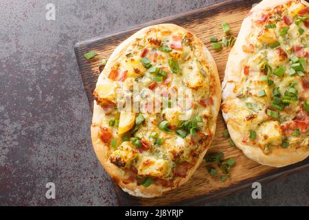 Offener Kuchen mit Käse, Kartoffeln, Zwiebeln und Speck aus der Nähe auf einem Holzbrett auf dem Tisch. Horizontale Draufsicht von oben Stockfoto