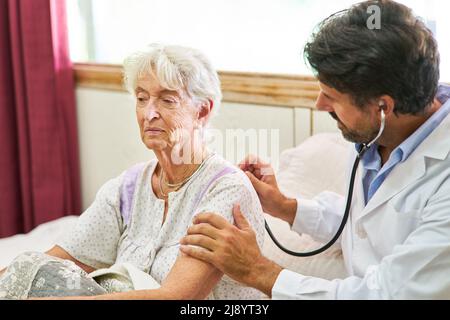 Kranke ältere Frau im Bett, die während des Hausbesuches mit Stethoskop vom Arzt untersucht wird Stockfoto
