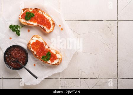 Sandwiches mit rotem Kaviar. Lachsroter Kaviar in der Schüssel und Sandwiches auf Marmorplatte auf altem, grau gesprenkeltem Tischhintergrund. Draufsicht. Speicherplatz kopieren. Stockfoto