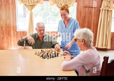 Ein altes Seniorenpaar im Pflegeheim spielt Schach neben einer lächelnden geriatrischen Krankenschwester Stockfoto