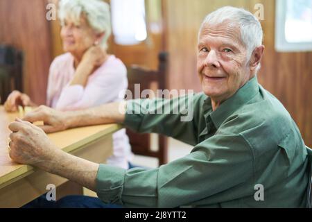 Senior mit Demenz sitzt am Tisch und spielt am Spielabend mit Freunden im Pflegeheim Dominosteine Stockfoto