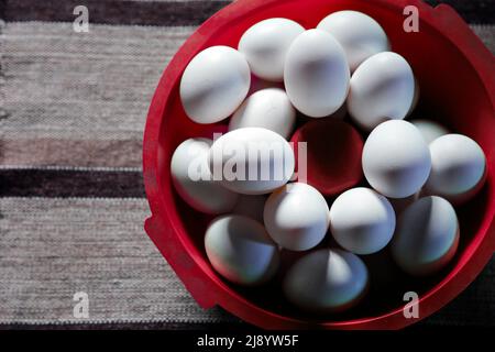 Verschiedene weiße Eier, gruppiert in einer roten Schüssel auf dem Tisch und weiche Seitenbeleuchtung Stockfoto