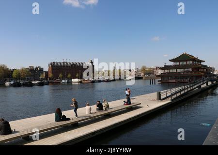Touristen sitzen und gehen auf dem schwimmenden Steg auf dem Kanal in Amsterdam in der Nähe des Hauptbahnhofs, Niederlande Stockfoto