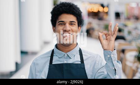 Junge afroamerikanische männliche Arbeiter Kellner Verkäufer in Schürze Mann Kleinbetrieb Inhaber des Cafés Restaurant Geschäft Blick auf die Kamera mit freundlichem Lächeln sh Stockfoto