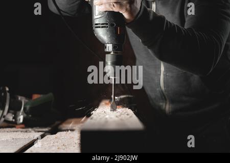 Ein Mann arbeitet mit einer Bohrmaschine in seiner Werkstatt. Zimmermann bohrt mit einem Hand-Elektrowerkzeug in einem dunklen Raum mit gerichtetem Licht Stockfoto