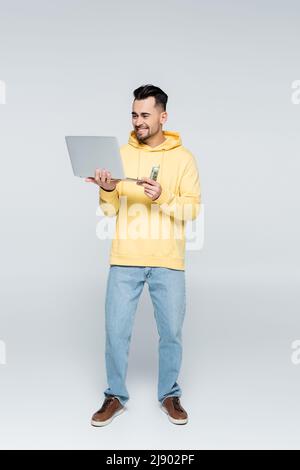 Volle Länge Ansicht von glücklichen Spieler mit Dollar und Laptop auf grau Stockfoto