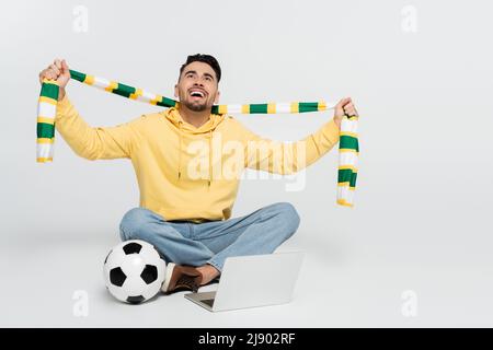 Fröhlicher Sportfan, der einen gestreiften Schal hält, während er in der Nähe des Laptops und des Fußballs auf Grau sitzt Stockfoto