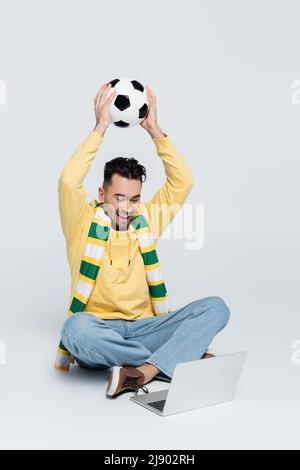 Fröhlicher Buchmacher mit Fußball sitzt mit gekreuzten Beinen in der Nähe von Laptop auf grau Stockfoto