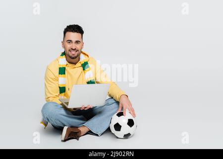 Zufrieden Spieler sitzt mit Laptop und Fußball, während Blick auf die Kamera auf grau Stockfoto