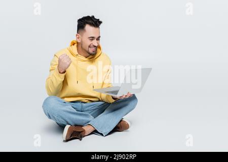 Aufgeregt Buchmacher zeigt Erfolg Geste in der Nähe Laptop, während auf grau sitzen Stockfoto