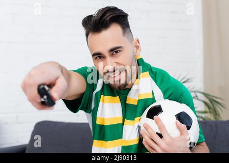 Lächelnder Sportfan mit Fußballball und verwackelte Fernbedienung, die zu Hause ein Spiel auf dem fernseher ansieht Stockfoto