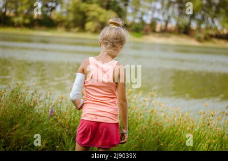 Kleines Kind mit Verbandszeug an gebrochener Hand, Rückenansicht in grüner Natur am Fluss Stockfoto