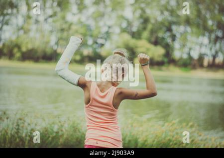 Kleines Kind mit Verbandszeug an gebrochener Hand, Rückenansicht in grüner Natur am Fluss Stockfoto