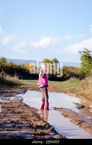 Kleines reizendes Mädchen, das nach dem Regen die Wasserpfütze in saisonaler herbstlicher, sonniger Atmosphäre genießt Stockfoto