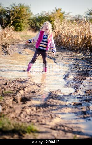 Kleines reizendes Mädchen, das nach dem Regen die Wasserpfütze in saisonaler herbstlicher, sonniger Atmosphäre genießt Stockfoto