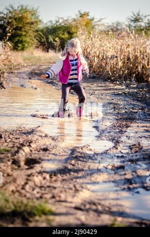 Kleines reizendes Mädchen, das nach dem Regen die Wasserpfütze in saisonaler herbstlicher, sonniger Atmosphäre genießt Stockfoto