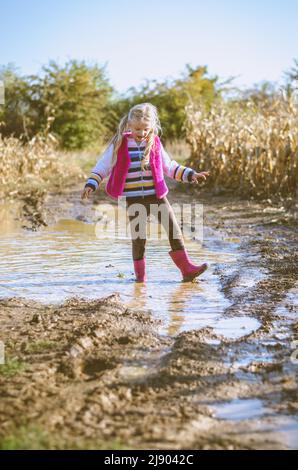 Kleines reizendes Mädchen, das nach dem Regen die Wasserpfütze in saisonaler herbstlicher, sonniger Atmosphäre genießt Stockfoto