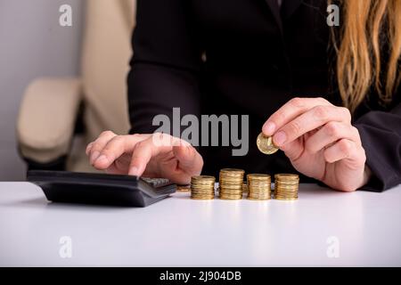 Eine Geschäftsfrau zählt auf einen Rechner und hält eine Münze in der Hand, an einem Tisch, vor Münzstapeln, aus der Nähe Stockfoto