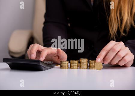 Eine Geschäftsfrau zählt auf einen Rechner und hält eine Münze in der Hand, an einem Tisch, vor Münzstapeln, aus der Nähe Stockfoto