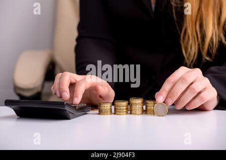 Eine Geschäftsfrau zählt auf einen Rechner und hält eine Münze in der Hand, an einem Tisch, vor Münzstapeln, aus der Nähe Stockfoto