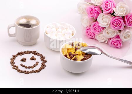 Gutes Frühstück. Müsli mit Milch, Kaffee mit Salmmilagen, ein Strauß Rosen und ein lächelndes Lächeln aus Kaffeebohnen Stockfoto