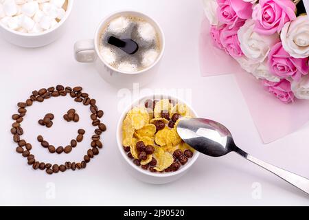 Gutes Frühstück. Müsli mit Milch, Kaffee mit Salmmilagen, ein Strauß Rosen und ein lächelndes Lächeln aus Kaffeebohnen Stockfoto